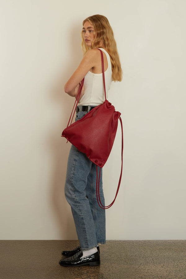 A woman wearing the red drawpack slung on her shoulder. The backpack has a long adjustable drawstring and a unisex design.