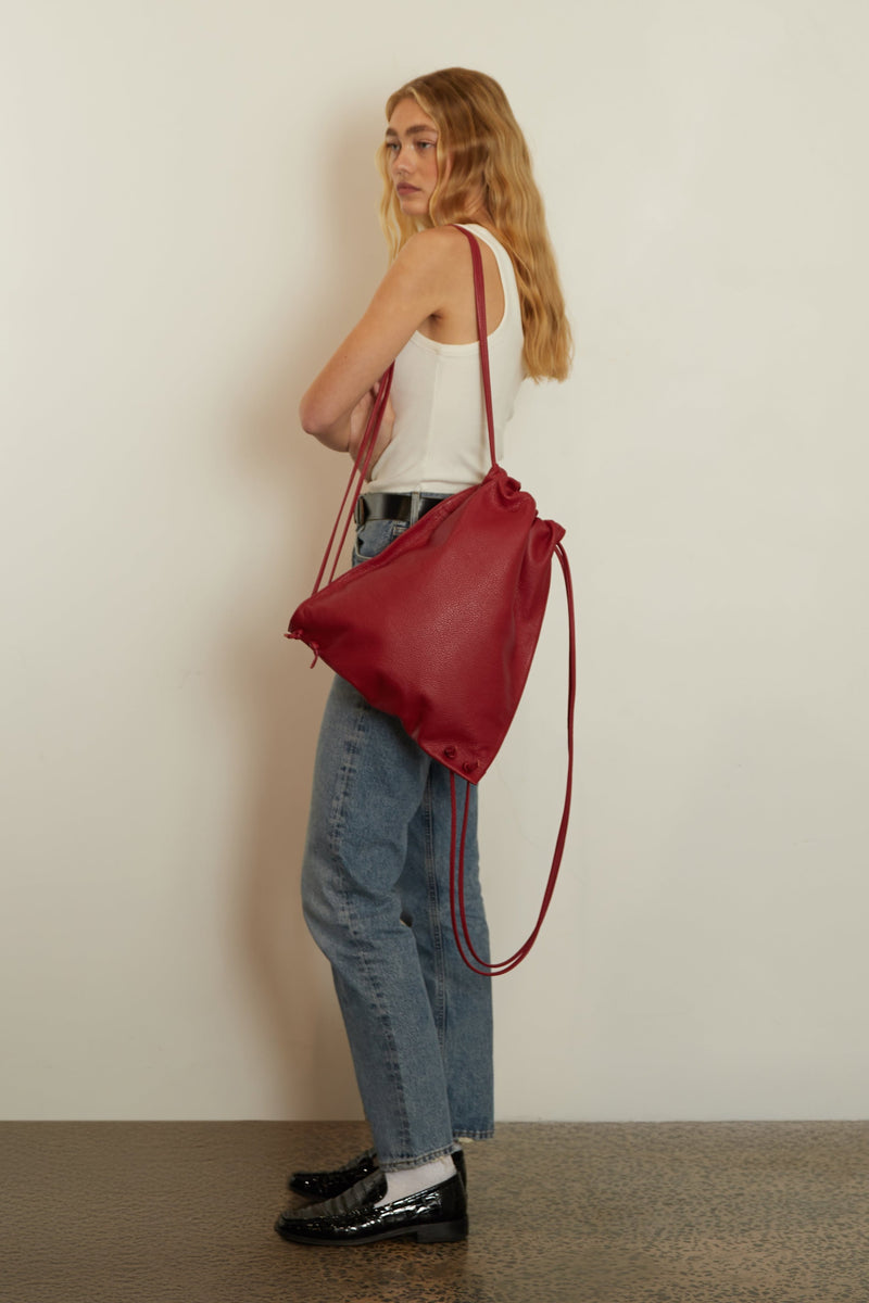 A woman wearing the red drawpack slung on her shoulder. The backpack has a long adjustable drawstring and a unisex design.