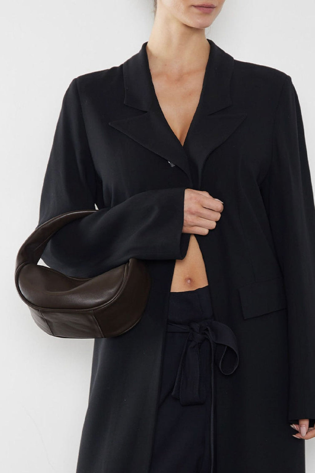 A woman wearing a the mini halo bag in the crook of the arm. It shows a front angle of the handbag showcasing its soft yet structured body. This clutch inspired mini bag is is suited as a handheld bag for parties and events.