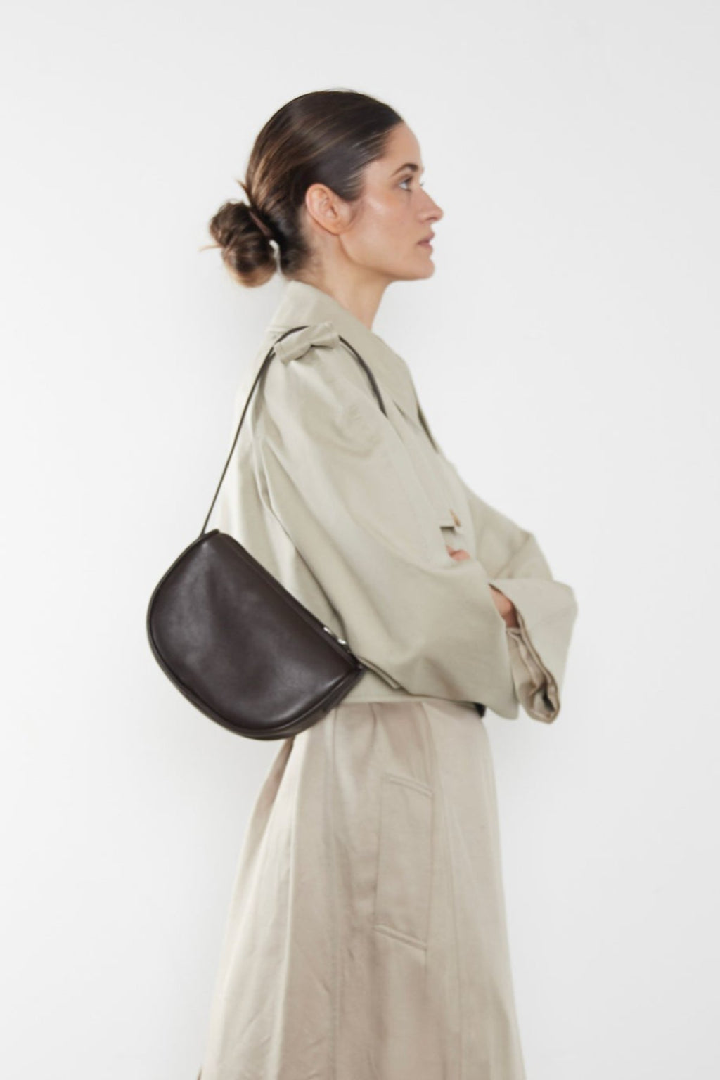 A woman carrying a dark brown mini shoulder bag. It shows a side angle of the saddle inspired bag showcasing its slim body. This small shoulder bag can carry everyday essentials such as sunglasses, wallet, car keys and lipstick