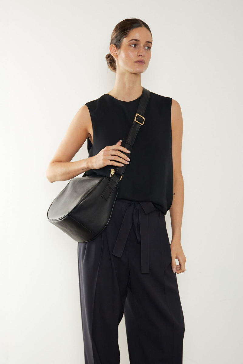 An image of a women carrying a black crossbody bag. The bag has a satchel like design. It is midsized with a structured body. The bag is called Saddle Crossbody Bag in Smooth Black.