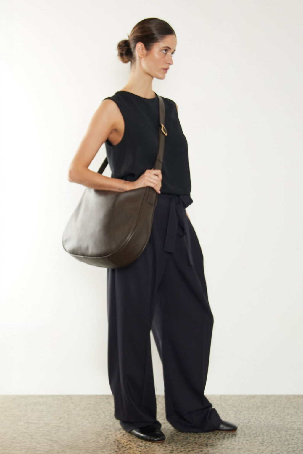 A woman wearing a crossbody bag slung across her body. It shows a side angle of the satchel showcasing its large but slim body. The bag is called Saddle Large Crossbody Bag in Smooth Dark Brown.