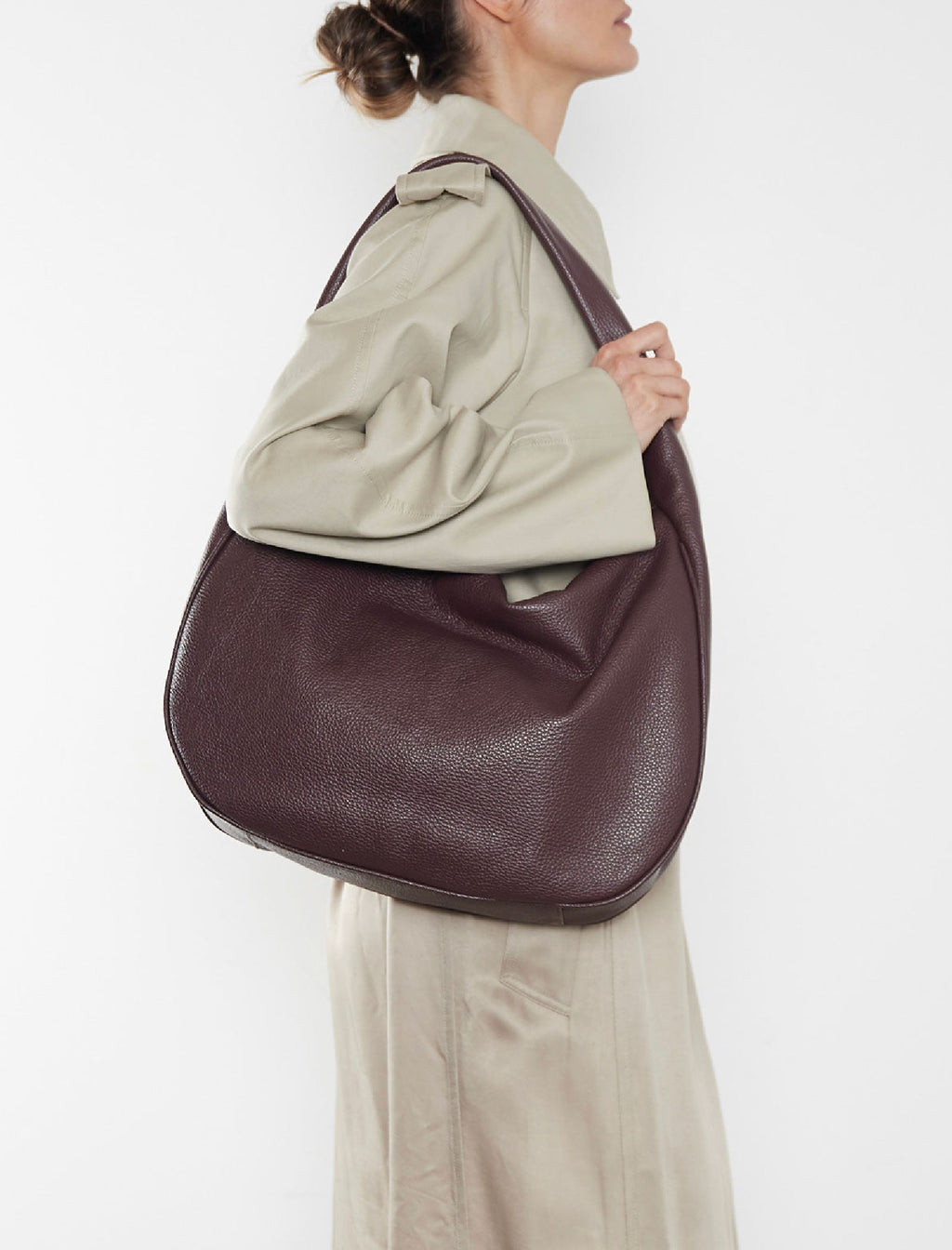 A woman wearing a burgundy shoulder bag. It shows a side angle of the satchel showcasing its large but slim body. It can carry everyday essentials and can be used as a work bag or a travel bag.