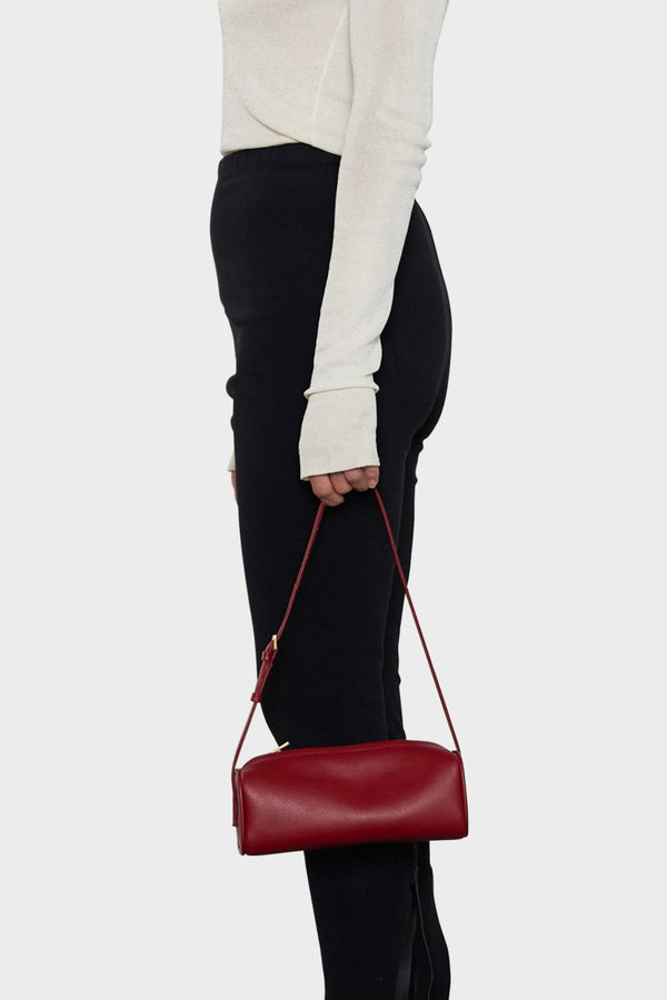 A woman holding the small red barrel shoulder bag in its handles. It highlights the elongated yet small and compact design of the handbag.