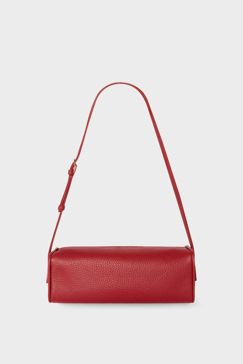 A small red shoulder bag made of grain Italian leather. The bag has a compact design and features an adjustable strap handle.